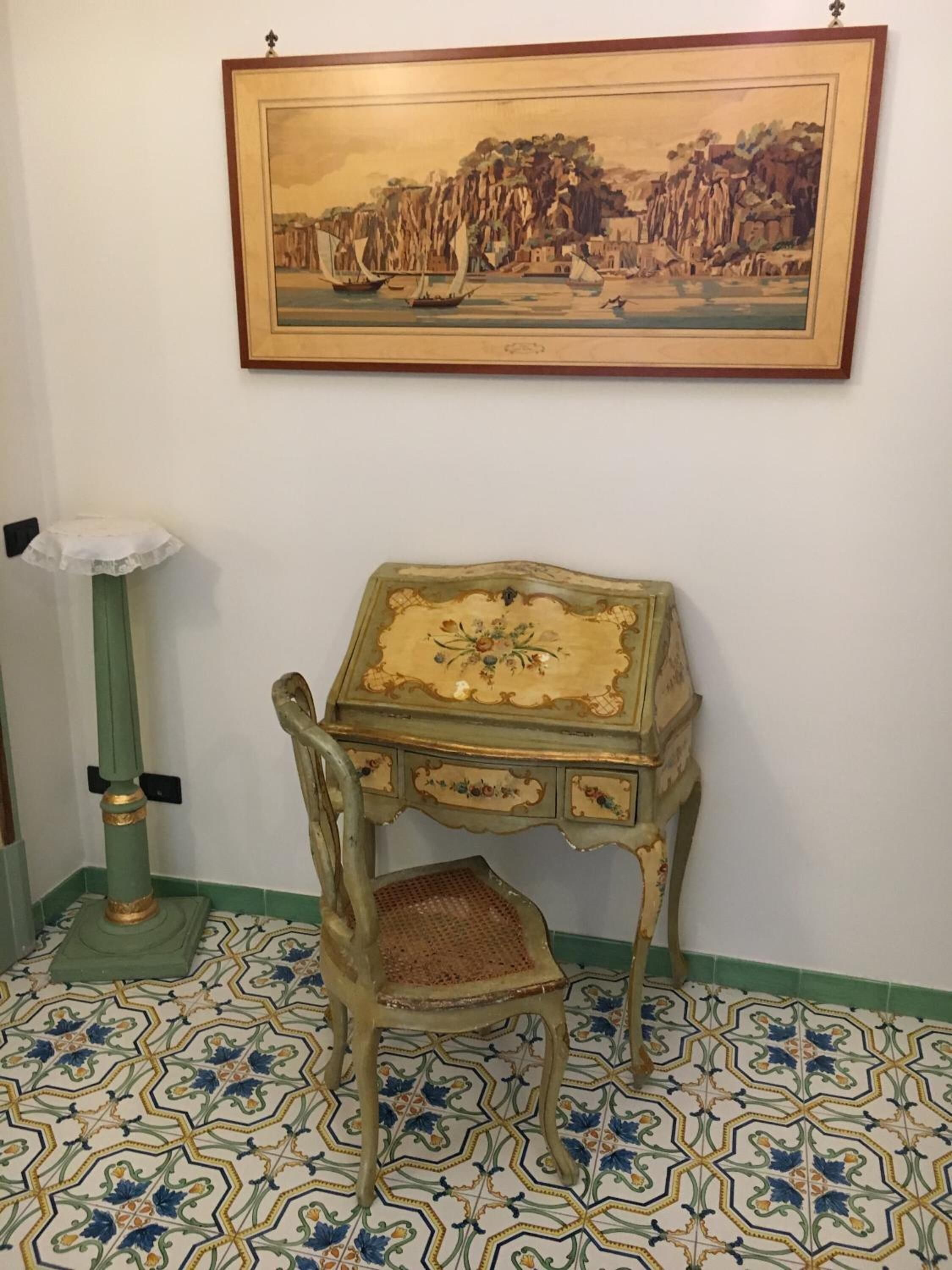 Ένα δωμάτιο με μια πράσινη καρέκλα, ένα μικρό τραπέζι με διακοσμητικά σχέδια λουλουδιών, έναν πίνακα που απεικονίζει μια παραθαλάσσια σκηνή και διακοσμητικά πλακάκια στο δάπεδο.