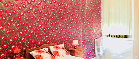 Quarto com papel de parede floral, cama de madeira, mesa de cabeceira, luminária e lustre.