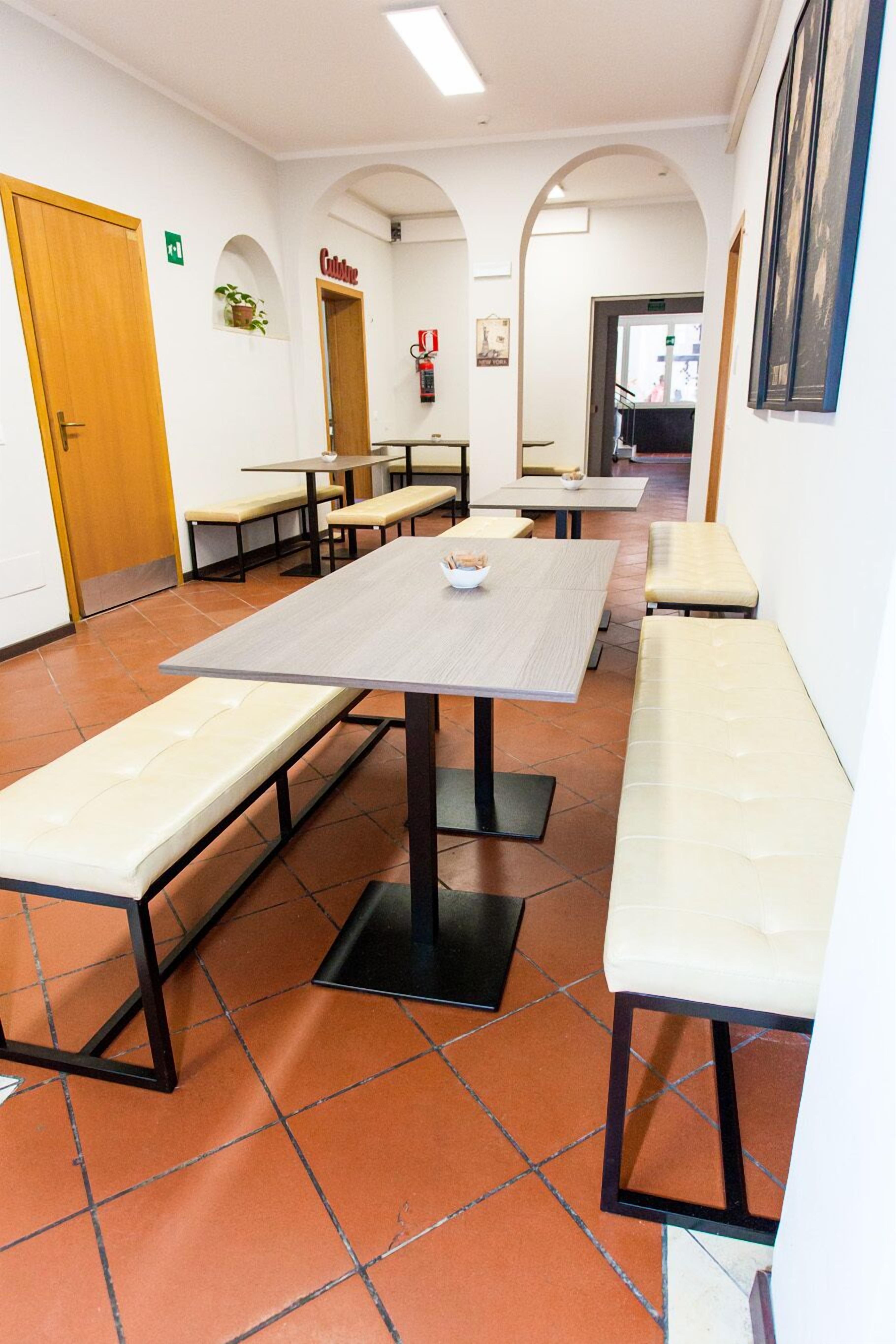 Un espace repas avec plusieurs tables et des bancs, un extincteur fixé au mur et une porte avec un panneau vert indiquant la sortie.