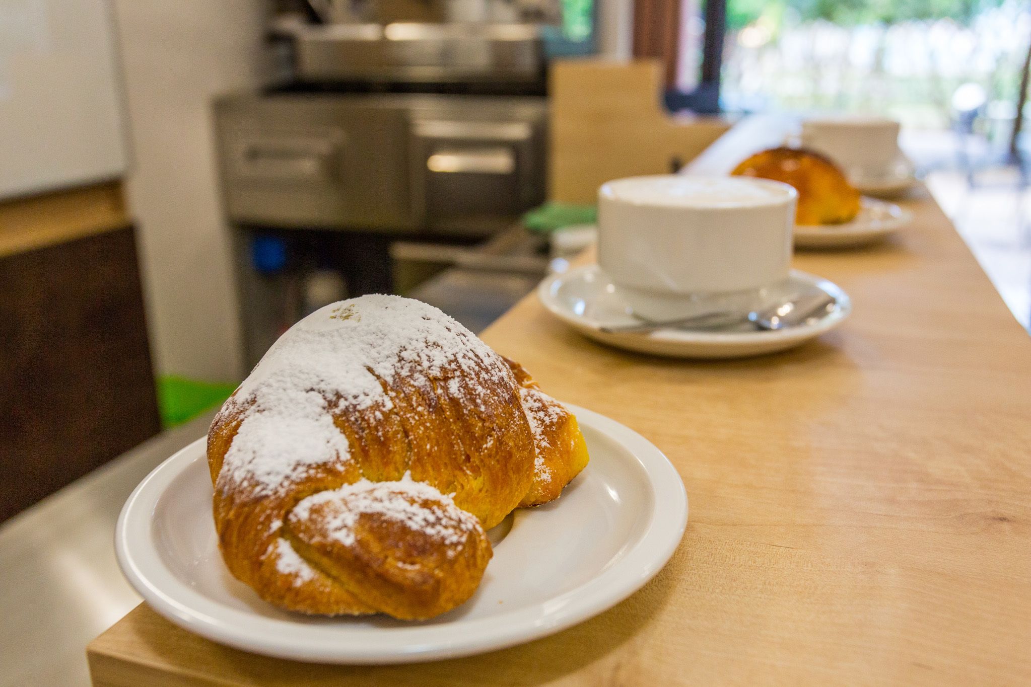 A croissant on a plate with powdered sugar, a cup of coffee on a saucer, and a pastry in the background.