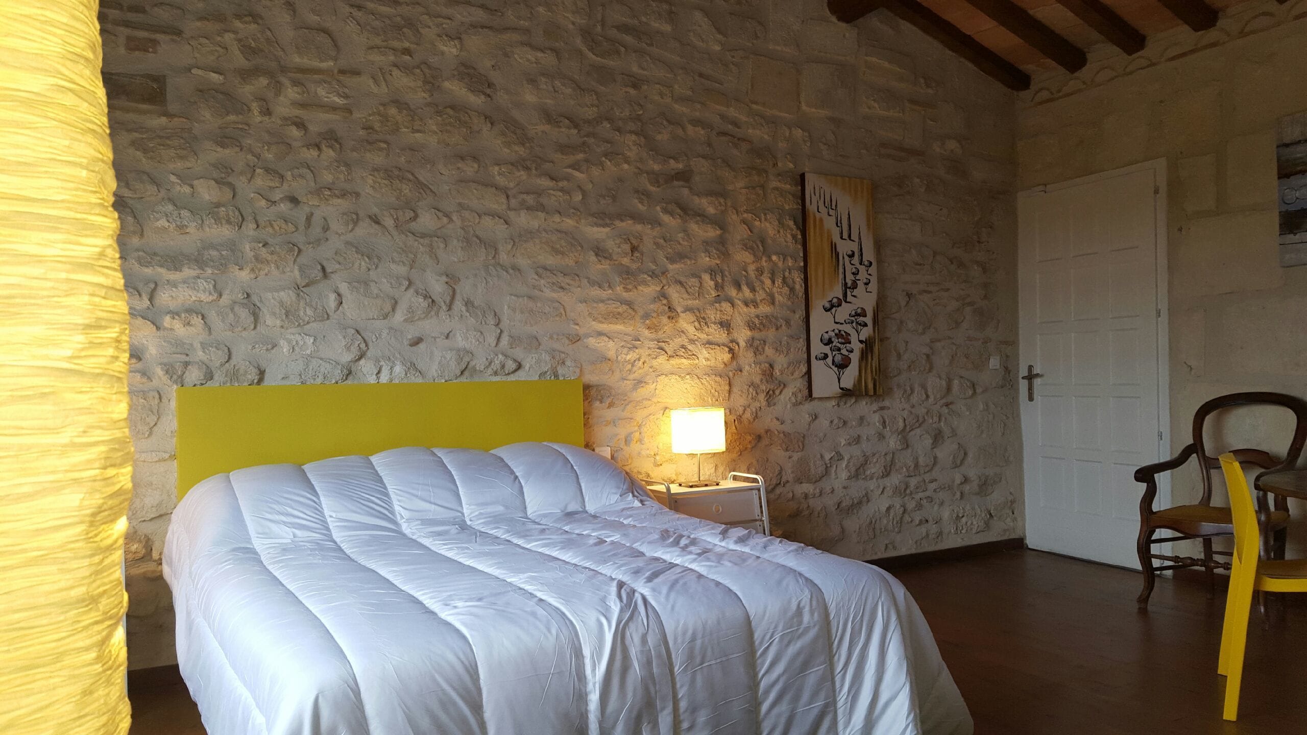Une chambre avec un mur en pierre, un lit avec une tête de lit jaune, une lampe de chevet, une chaise en bois et une porte.