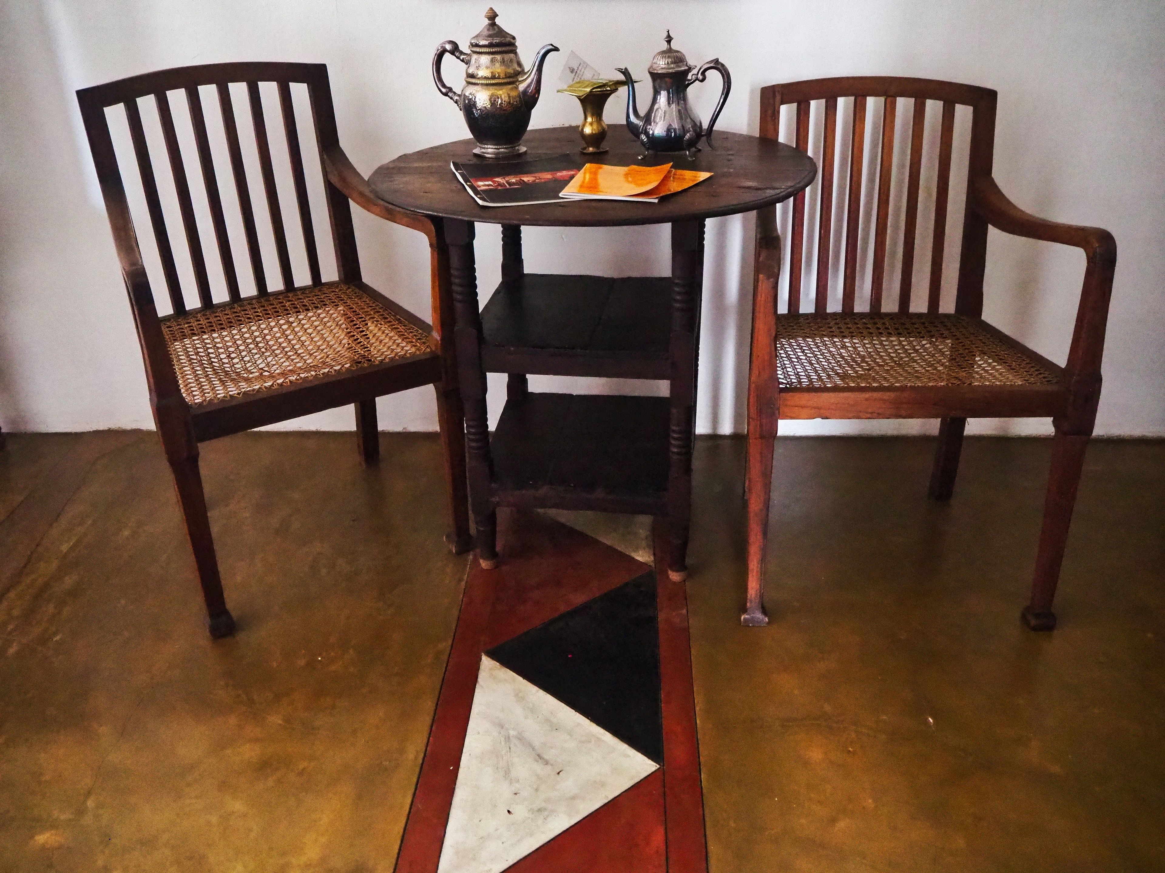 Een ronde houten tafel met twee stoelen, elk met een geweven zitting. Op de tafel staan twee metalen theepotten en een boek.