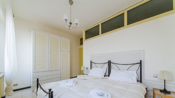 A neatly made bed with white linens, a bedside table with a lamp, and a window with blinds. - Welc-oM Prato della Valle (Padova)