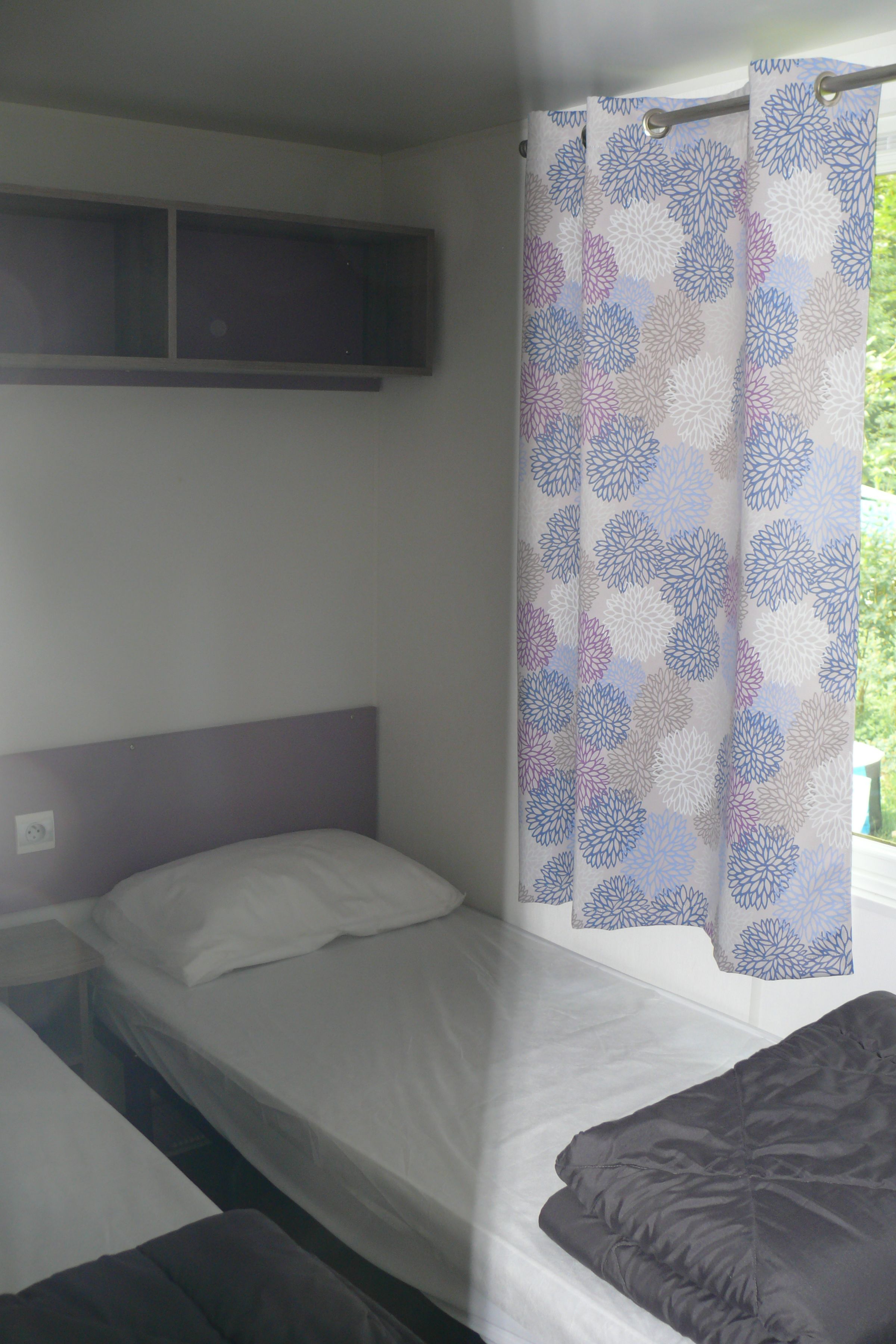 Un lit simple avec une tête de lit, une fenêtre avec un rideau et une petite étagère au-dessus du lit.