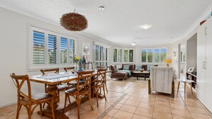 A spacious living area with a dining table, chairs, and a sofa. There are large windows with shutters, a ceiling fan, and a hanging light fixture. - SHORELINE 3 BEACH FRONT APARTMENT (HASTINGS POINT)
