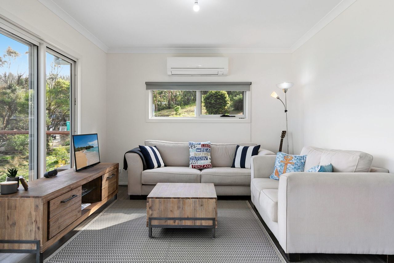 A modern living room with a sofa, a coffee table, a TV on a wooden cabinet, and a window with a view of greenery.