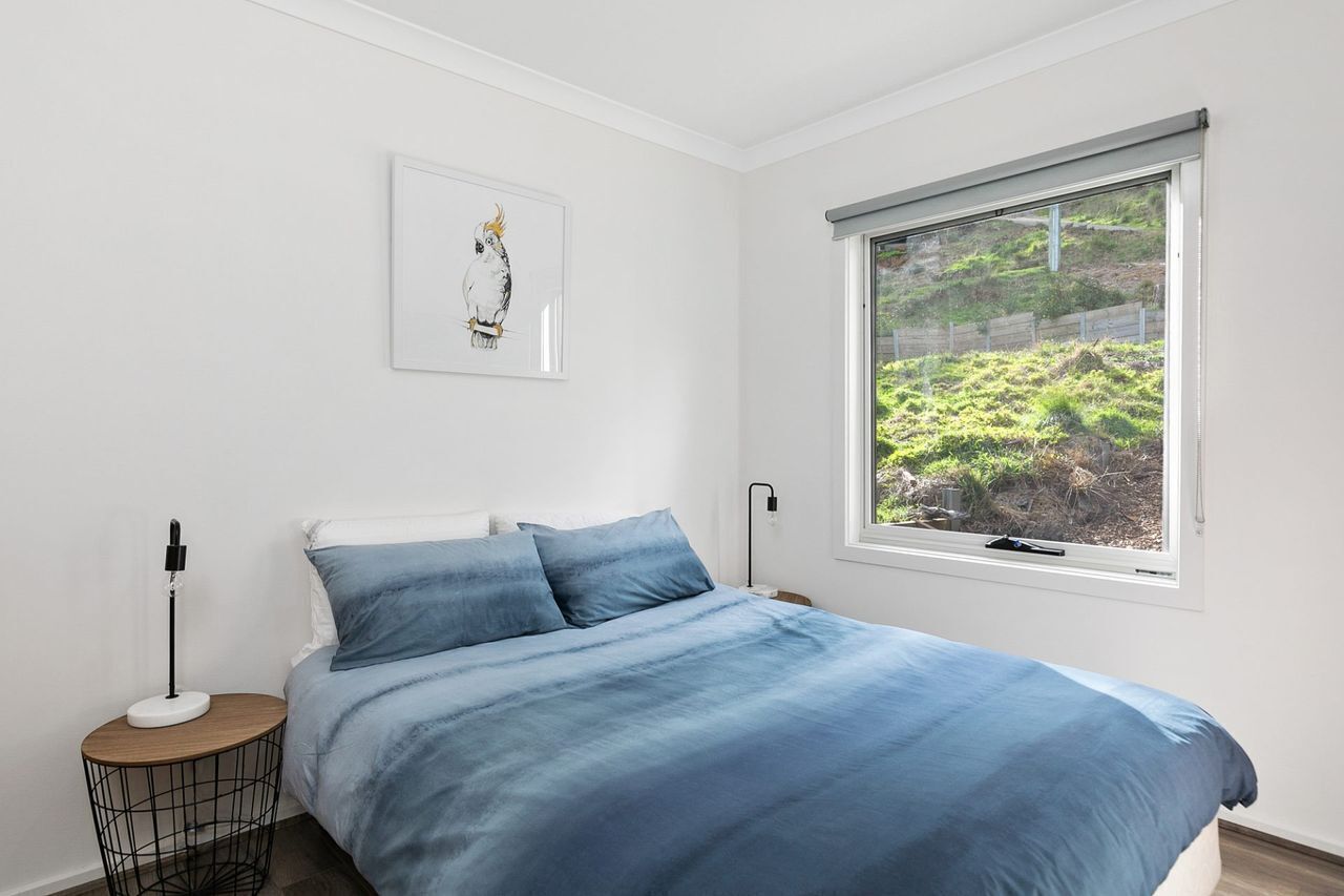 Un dormitorio con cama, mesita de noche, lámpara, ventana con vistas a vegetación y un cuadro en la pared.