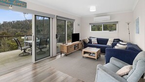 Ein modernes Wohnzimmer mit einer Couch, einem Couchtisch und einem Fernseher. Eine Glastür öffnet sich zu einem Balkon mit Gartenmöbeln.