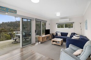 A modern living room with a sofa, coffee table, and a TV. There is a sliding glass door leading to a balcony with outdoor furniture. - Hull's Haven (Wye River)