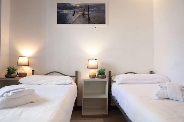 A hotel room with two beds, each with white linens and pillows, a small bedside table with a lamp and a plant, and a wall-mounted picture of a pier.