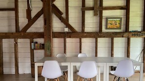 Comedor con una mesa larga y cuatro sillas blancas de patas de madera. La habitación tiene vigas de madera a la vista y una pared de ladrillo blanco.
