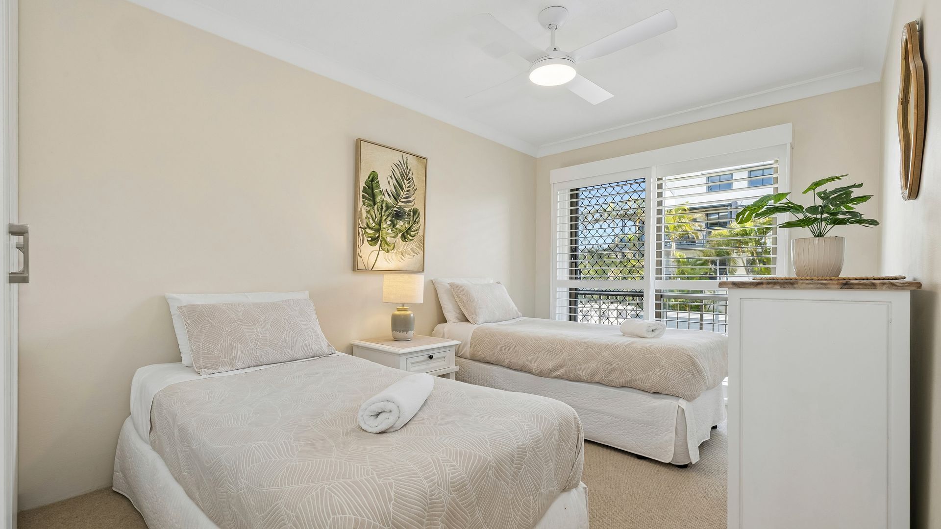 A bedroom with two beds, a ceiling fan, a window with a view of greenery, and a painting on the wall.
