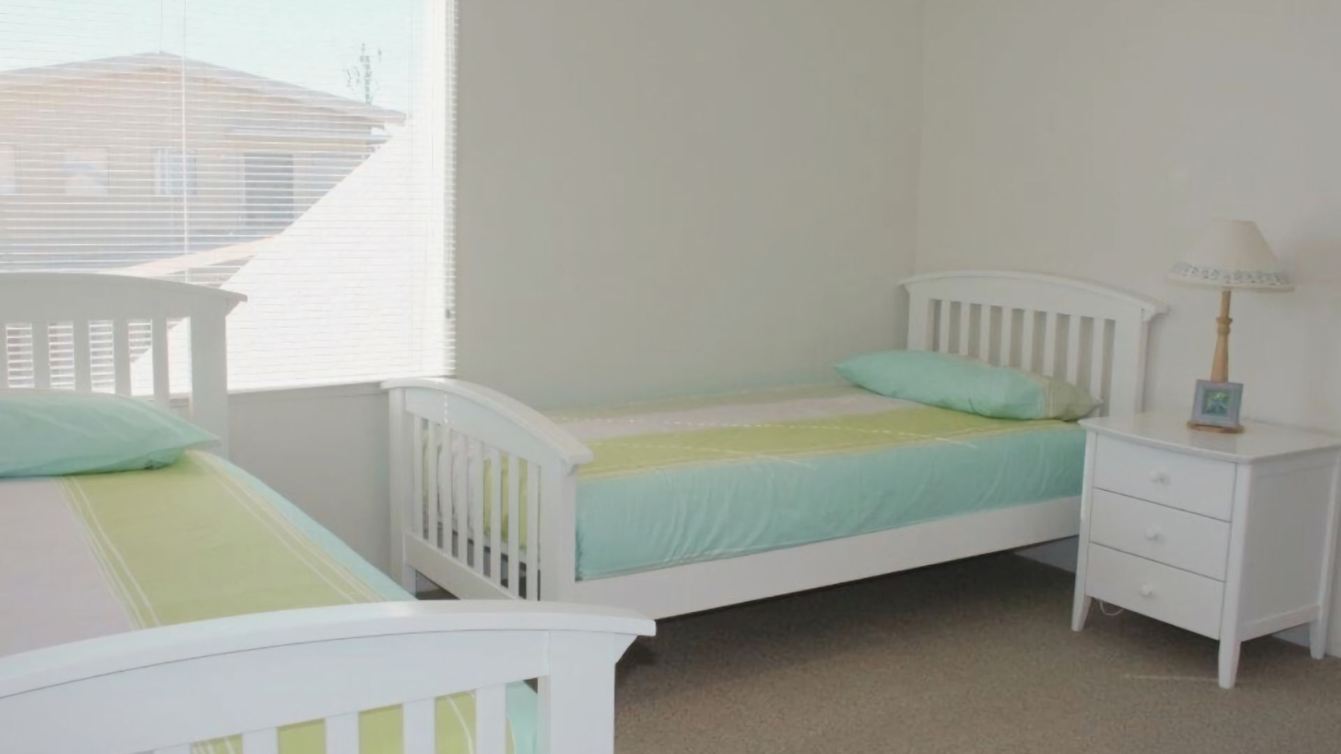 A bedroom with two white beds, a nightstand with a lamp, and a window with blinds.