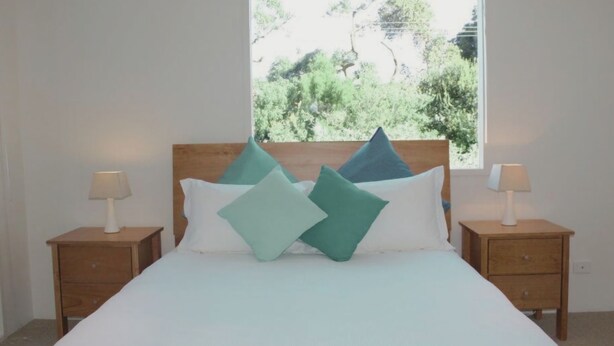 A bed with a wooden headboard, two bedside tables with lamps, and a window with a view of greenery. - Lonsdale Shores in Point Lonsdale (Point Lonsdale)
