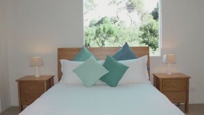 A bed with a wooden headboard, two bedside tables with lamps, and a window with a view of greenery. - Lonsdale Shores in Point Lonsdale (Point Lonsdale)