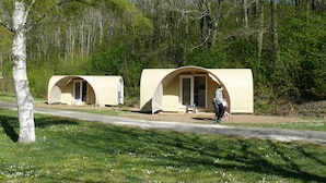 Zwei kleine, beige Zelthaus-ähnliche Gebilde mit Türen, eingebettet in einen bewaldeten Bereich mit einem Feldweg, der zu ihnen führt.