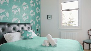 A bedroom with a tufted headboard, a bed with a green bedspread, two pillows, a rolled-up towel, a framed picture on the wall, and a window with a view of trees. - Ozone Tower in Queenscliff (Queenscliffe)