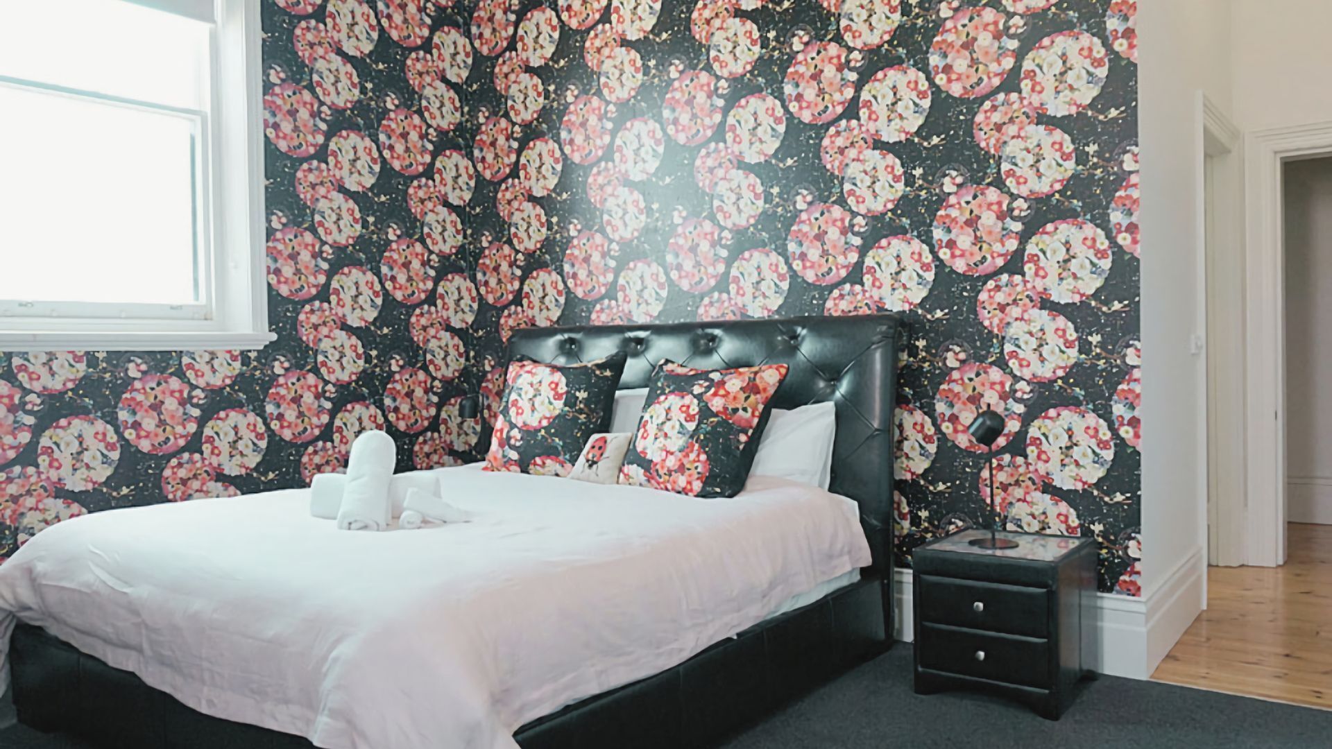 A bedroom with a floral wallpaper, a black upholstered headboard, a white bedspread, and a bedside table with a lamp.