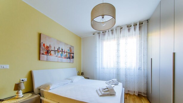 Ein Schlafzimmer mit Bett, Nachttisch, Lampe, einem Bild, einem Fenster mit Vorhängen und einem Kleiderschrank.