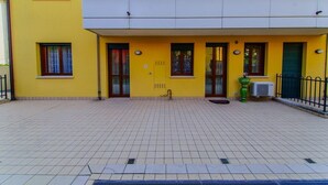 Ein gelbes Gebäude mit flachem Dach, vielen Fenstern und einer gefliesten Fläche vor der Fassade.