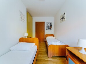 A small, clean room with two single beds, a wooden wardrobe, a desk with a lamp, and a door leading to another room.