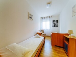 A bedroom with a single bed, a wooden desk, a chair, and a small table with a lamp.