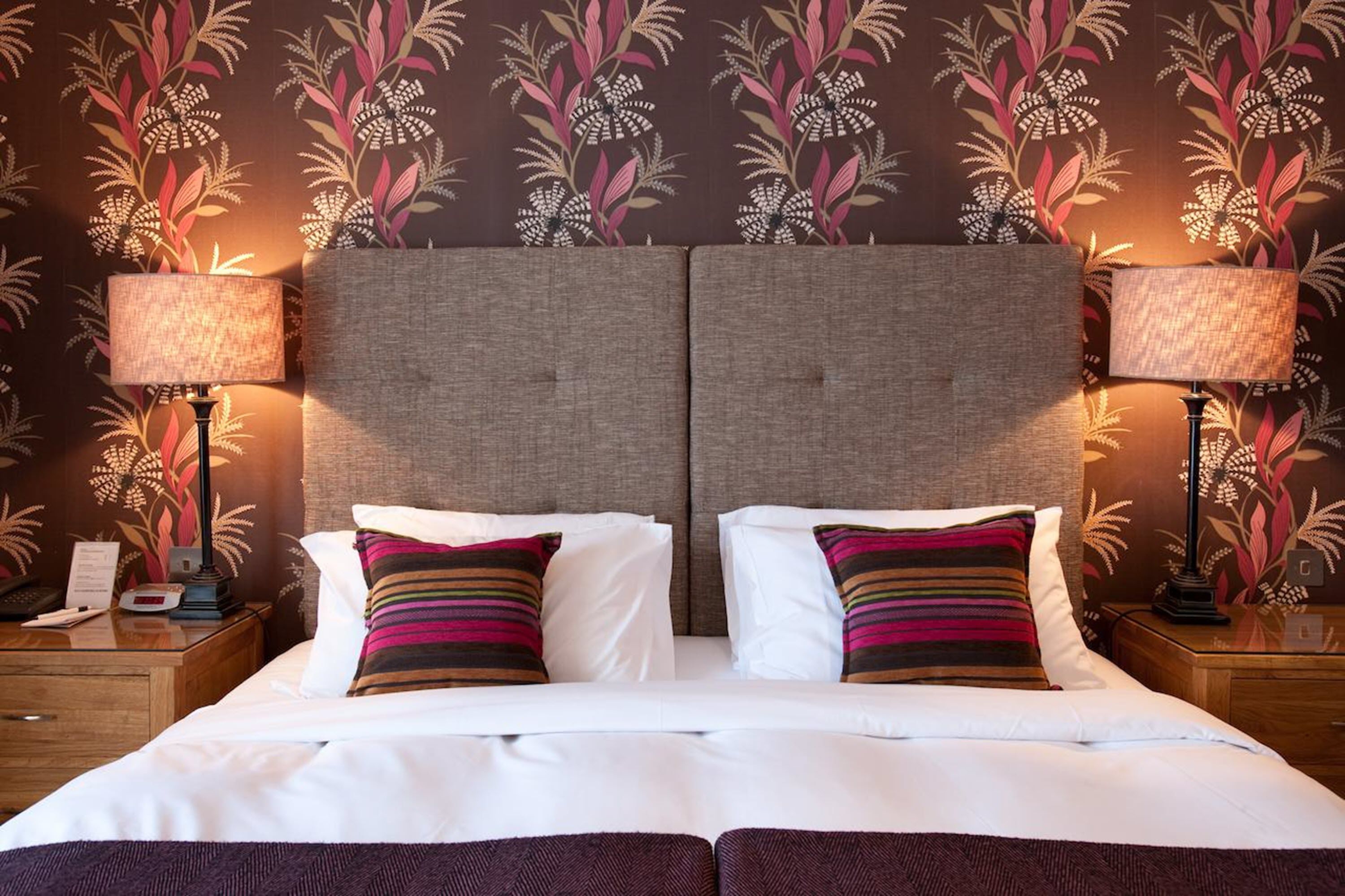 A double bed with white linens and two pillows, flanked by two nightstands with lamps, against a wall with a floral wallpaper pattern.