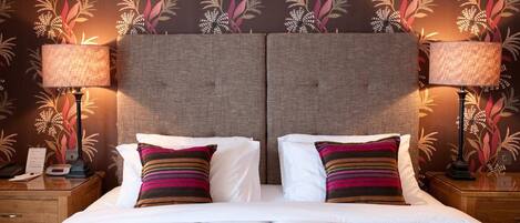 A double bed with white linens and two pillows, flanked by two nightstands with lamps, against a wall with a floral wallpaper pattern.
