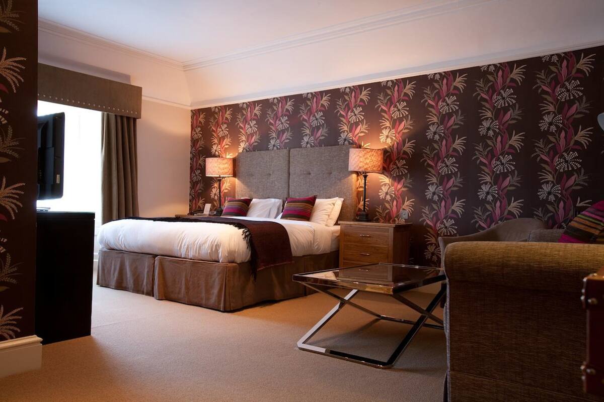 A hotel room with a bed, bedside lamps, a glass coffee table, a chair, and a dresser.