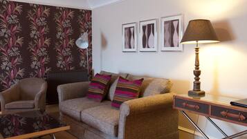 Un salon avec un canapé, un fauteuil et une petite table. Il y a trois tableaux encadrés au mur, une lampe sur une table d’appoint et un radiateur.