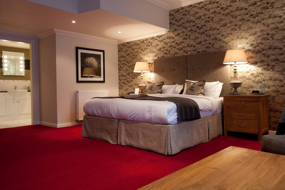 A bedroom with a large bed, two bedside lamps, a wooden nightstand, a red carpet, a radiator, a framed picture on the wall, and a bathroom visible through an open door.