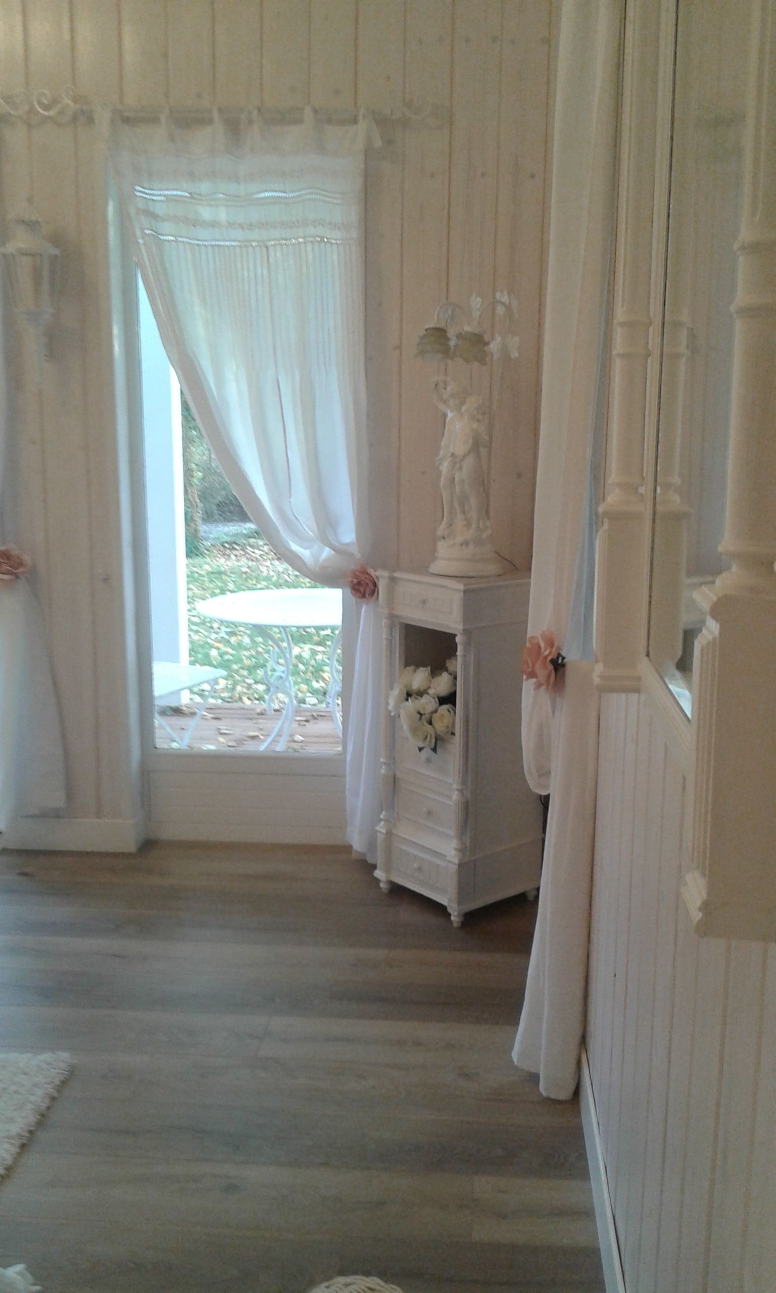 Una habitación con piso de madera, un tocador blanco con una estatua y flores, y una ventana con cortinas blancas.