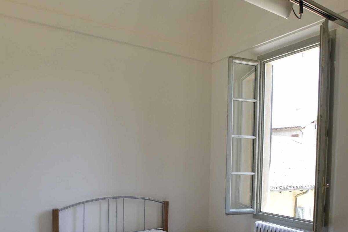 A bedroom with a single bed, a nightstand, a chair, and a window with an open shutter.
