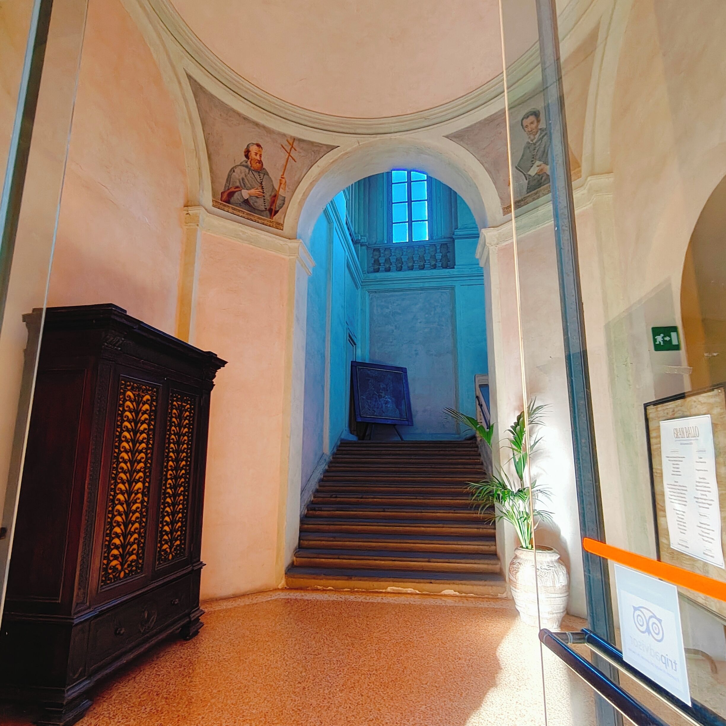 A hallway with a staircase leading to a blue-lit room, a dark wooden cabinet, and a framed certificate on a stand.