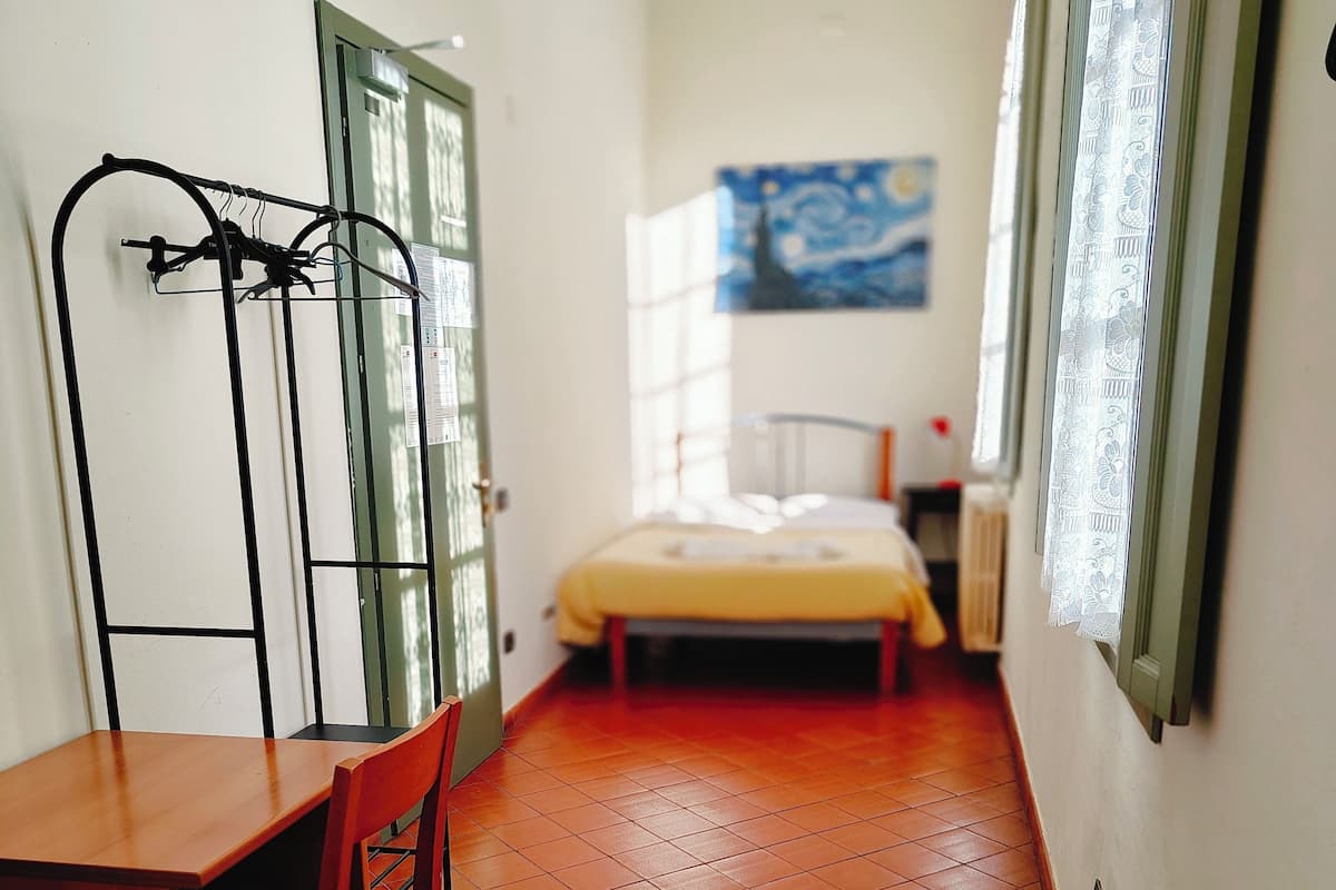 A small, narrow room with a wooden desk and chair, a bed with a yellow bedspread, and a painting on the wall.