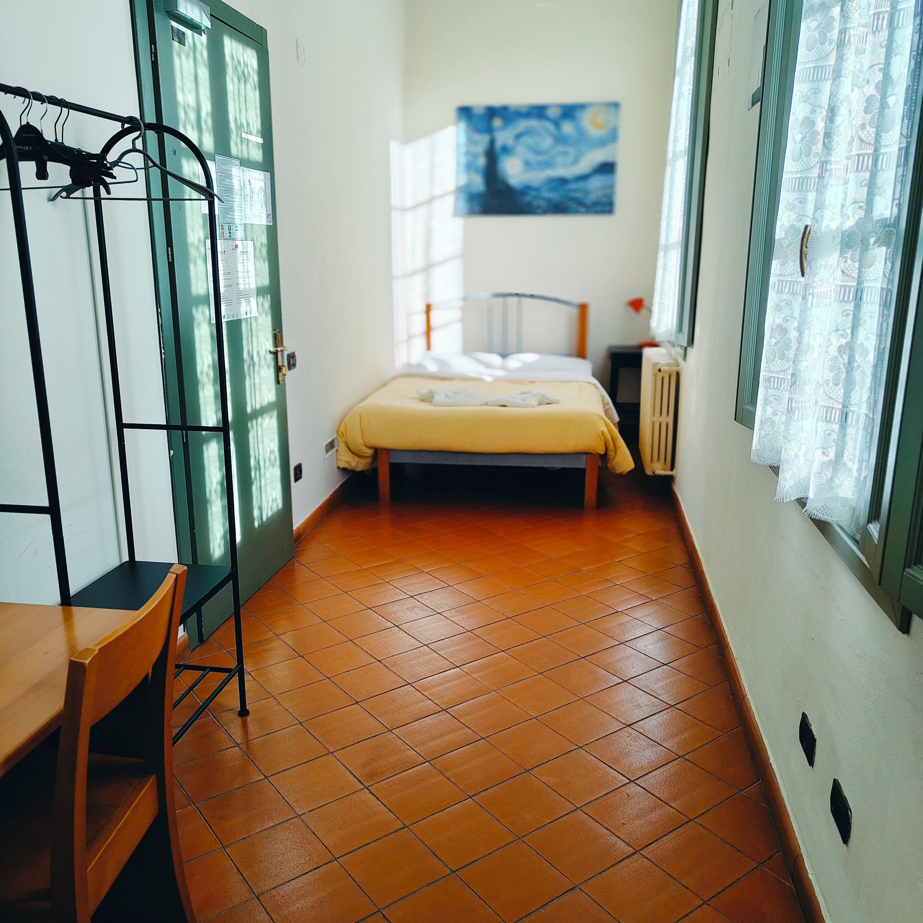 A narrow room with a bed, a wooden chair, a table, and a radiator.