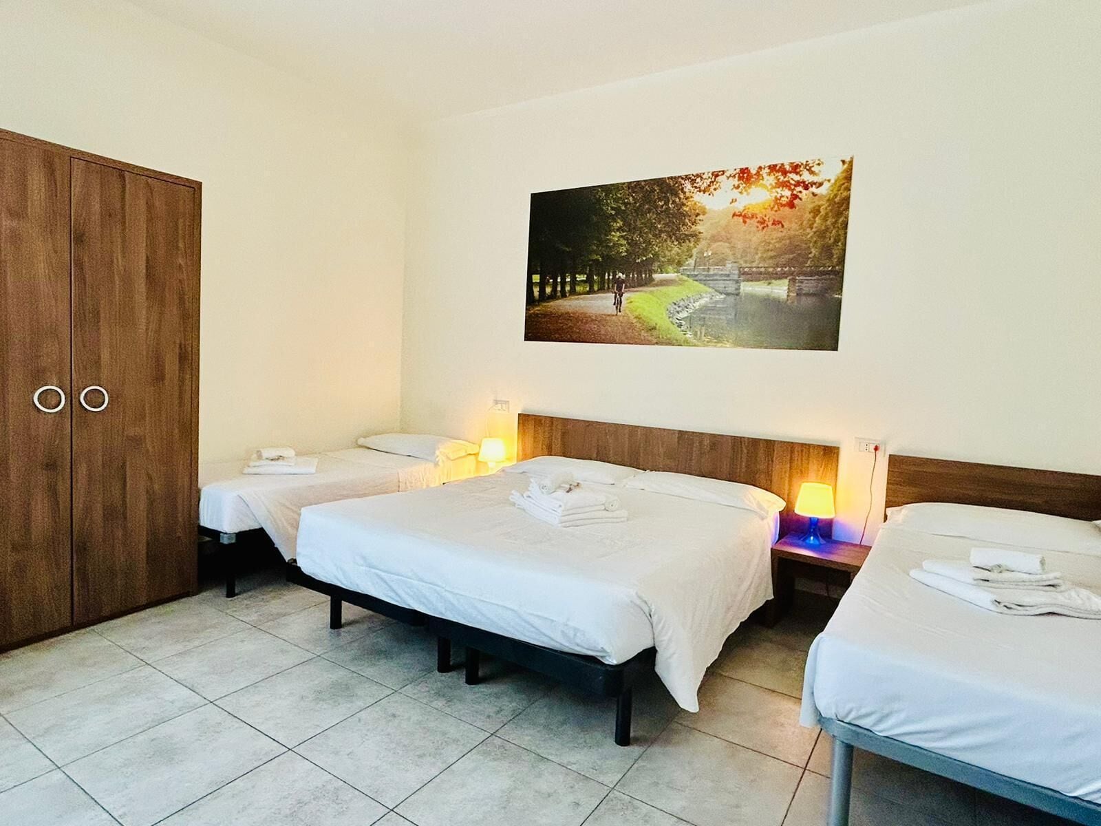 Une chambre d’hôtel avec trois lits simples, une armoire en bois et un tableau représentant un paysage pittoresque accroché au mur.