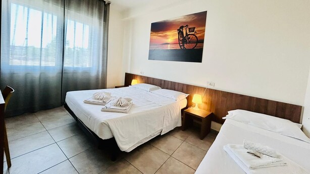 Una habitación de hotel con cama, mesita de noche con lámpara, una silla y ventana con cortinas.