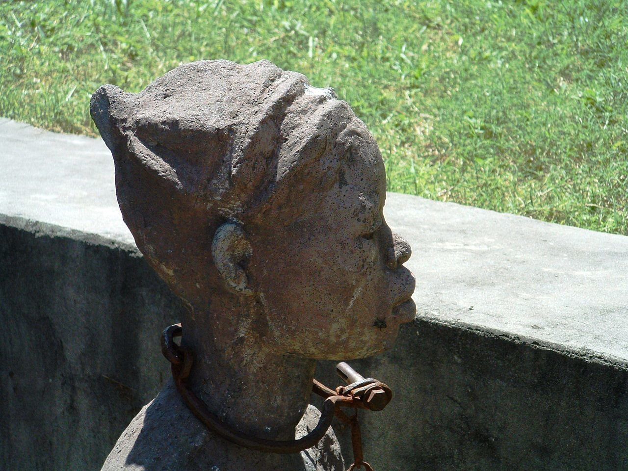 A weathered stone bust with a chain, set against a concrete wall with grass in the background.