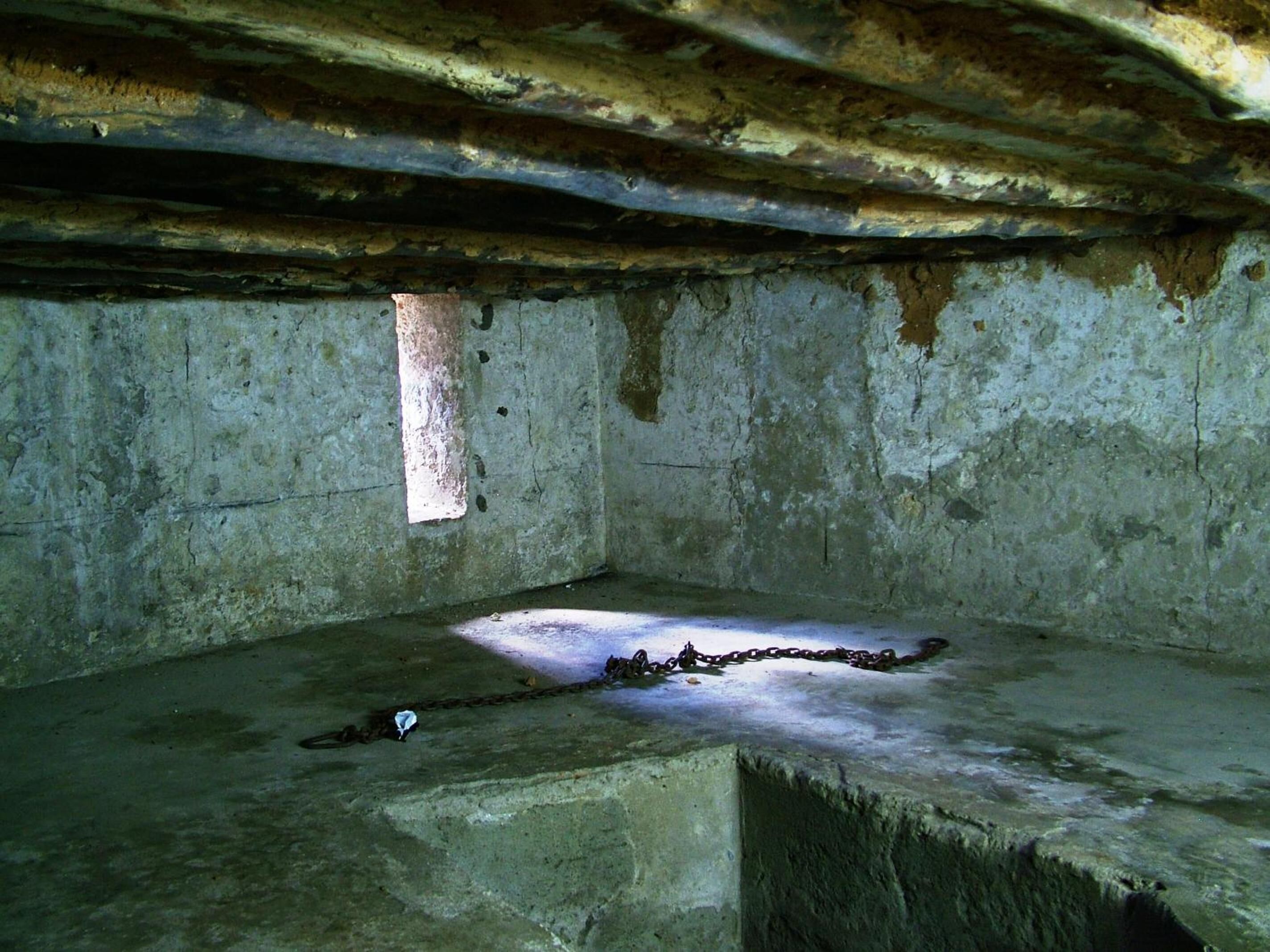 A dimly lit, confined space with concrete walls and a slanted ceiling, featuring a small window and a chain on the floor.