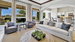 A spacious living room with a large window, a sofa, a coffee table with plants, and a kitchen in the background. - Stunning PGA  Home with Privacy and Lake Views on Golf Course (La Quinta)