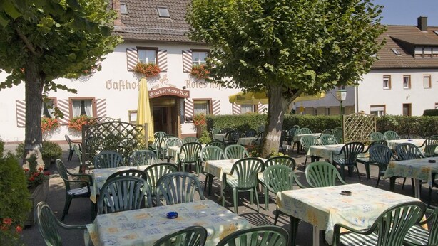 An outdoor seating area with tables and chairs, a building with a sign reading 'Gasthof Rotes Röß', and a tree in the background.