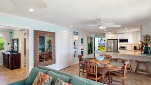 A spacious living area with a green sofa, wooden dining table, and chairs. There is a kitchen with white cabinets and a refrigerator.