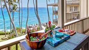 Un balcon avec vue sur l’océan, un saladier en bois rempli de salade et une table dressée avec des assiettes et des verres.