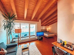 En stue med fjernsyn, sofa, sofabord, en plante og en balkon med udsigt.