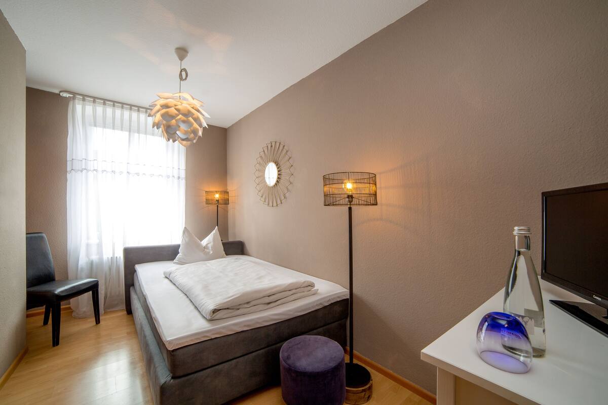Ein Schlafzimmer mit Bett, Nachttischlampen, einem Schreibtisch mit Computer und einem Fenster mit Vorhängen.