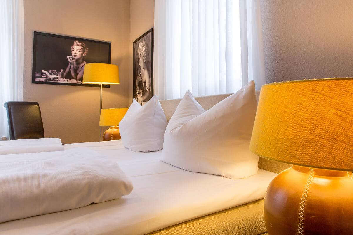 Ein Hotelzimmer mit einem ordentlich bezogenen Bett, einem großen Fenster mit Vorhängen, einem an der Wand montierten Fernseher und einer Lampe.