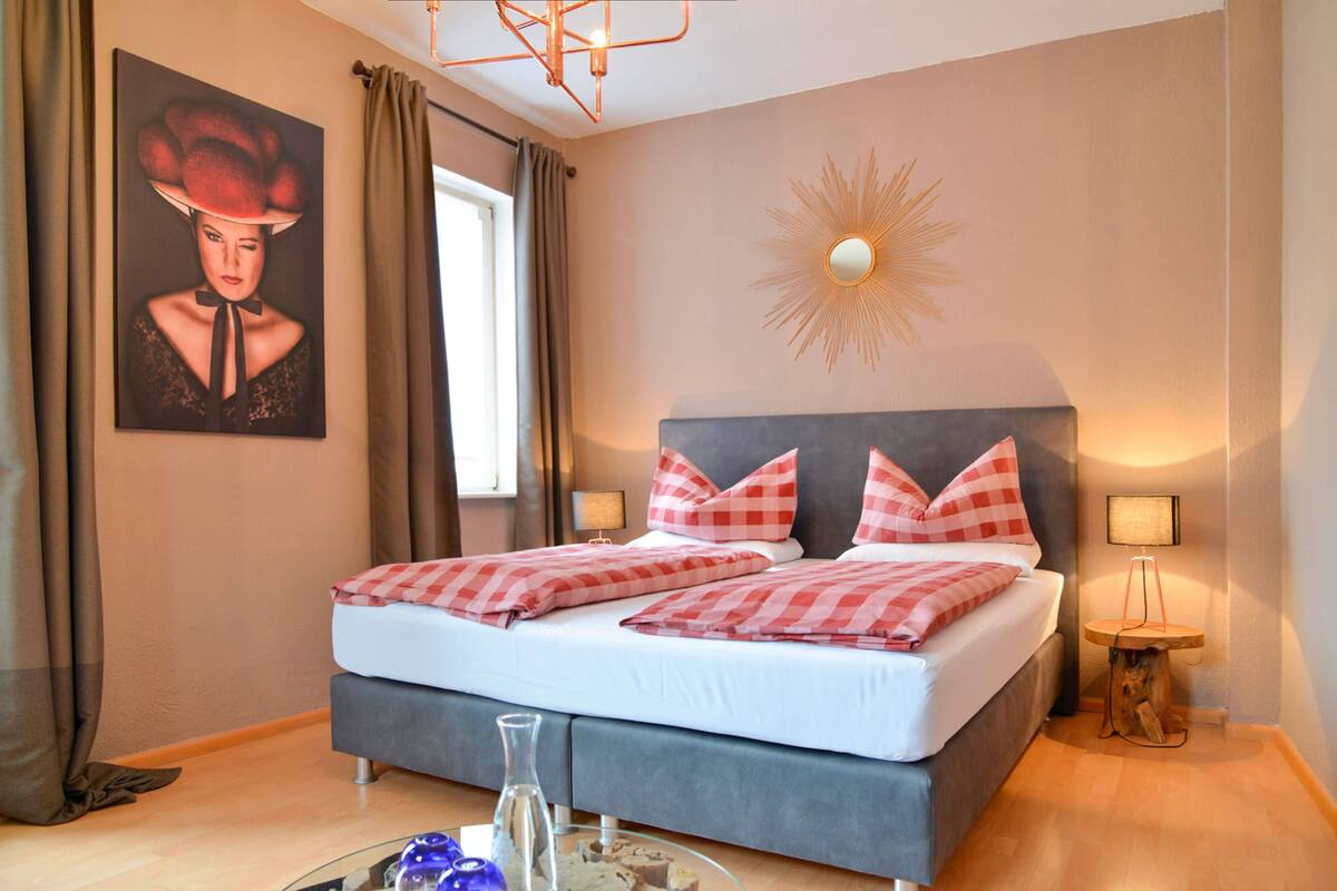 Ein Schlafzimmer mit einem Bett, Karokissen, einer Wanddekoration im Sonnenstrahlendesign, einem Nachttisch mit Lampe und einem Bild an der Wand.