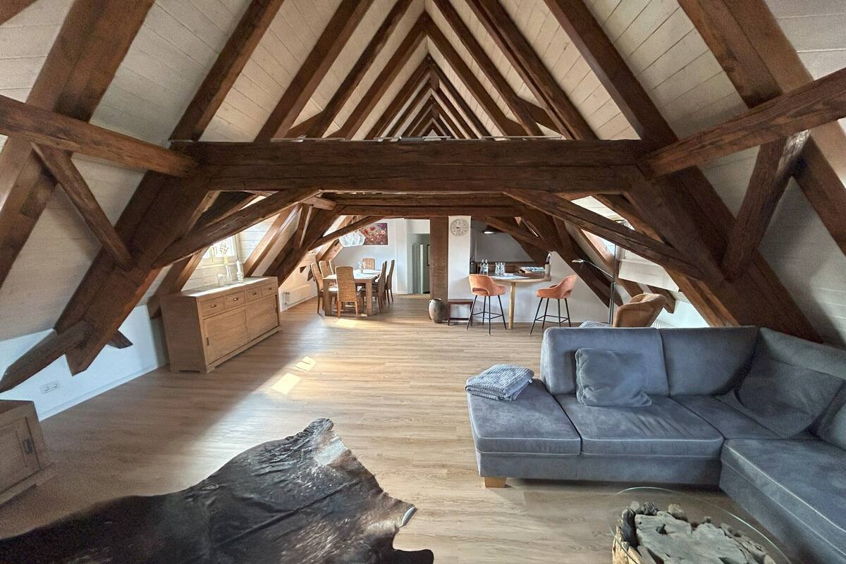 Ein Dachgeschossraum mit freiliegenden Holzbalken, einem Essbereich mit rundem Tisch und Stühlen, einer Couch und einem Couchtisch.
