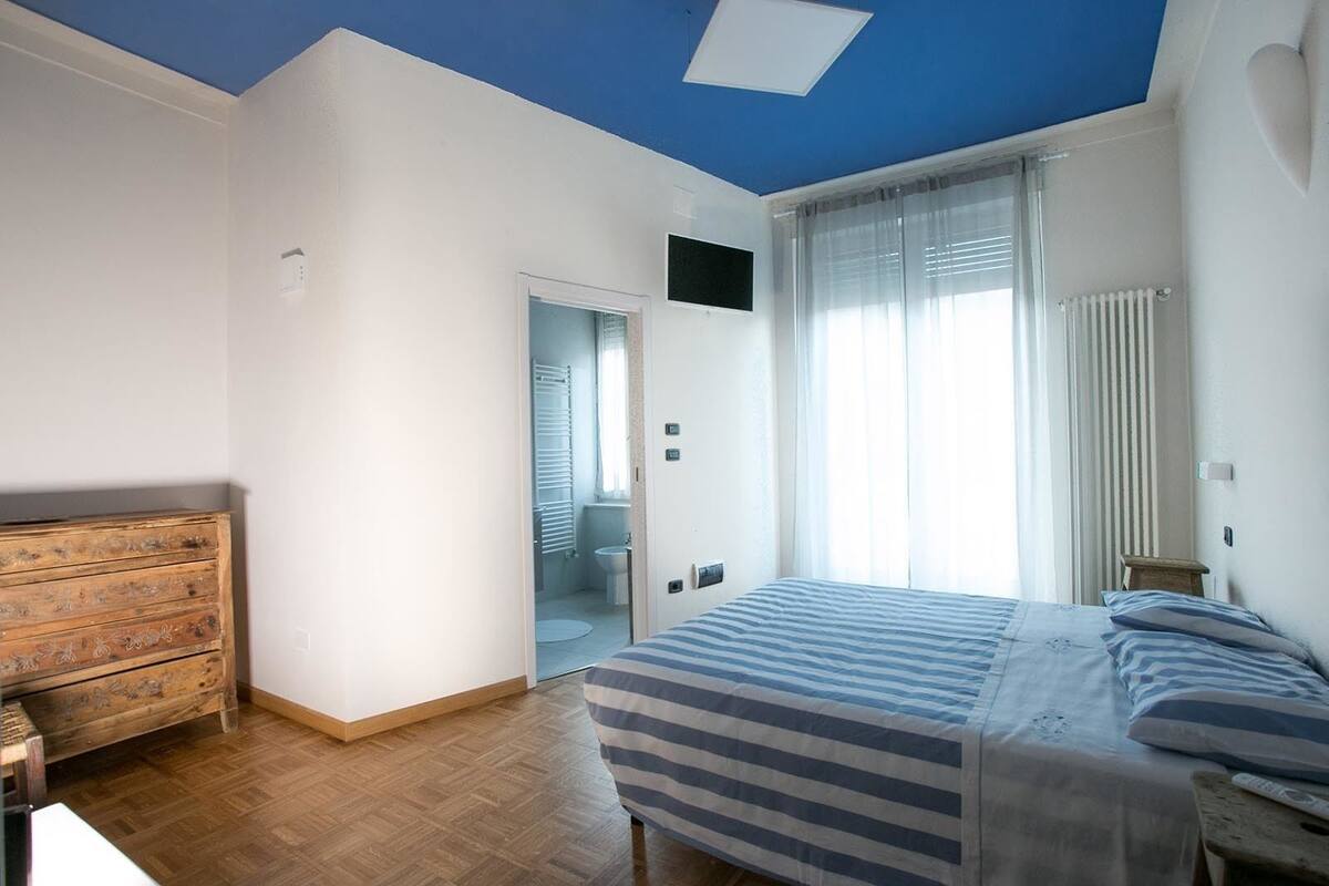 A bedroom with a bed, a wooden dresser, a bathroom with a shower and toilet, and a ceiling light fixture.